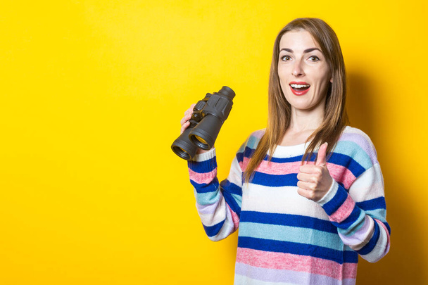 Молода жінка з посмішкою робить привітний жест і тримає бінокль в руках на жовтому тлі. Банер. - Фото, зображення
