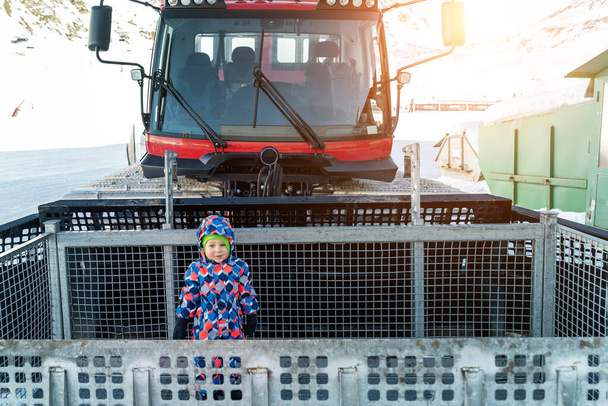 Χαριτωμένο αξιολάτρευτο παιχνιδιάρικο χαρούμενος αγοράκι μέσα στο κόκκινο σύγχρονο snowcat ratrack snowplow box περιποίηση στέκεται στην κορυφή αλπικό θέρετρο σκι Ischgl Αυστρία. Βαρέα μηχανήματα οχημάτων τροχιάς εξοπλισμού βουνού - Φωτογραφία, εικόνα