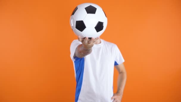 Ventilador de fútbol en camisa blanca azul de pie y sosteniendo la pelota delante de su cara con una mano - Metraje, vídeo