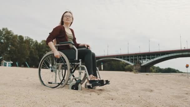 Tekerlekli sandalyedeki genç kadın kumlu nehir kıyısında sonbahar rüzgarının tadını çıkarıyor. - Video, Çekim