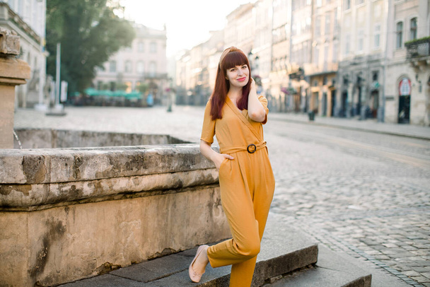 Charming young Caucasian dark haired woman, wearing elegant fashionable yellow clothes, smiling to camera while standing on ancient city street on old stone fountain. Outdoor city shot. - Fotografie, Obrázek