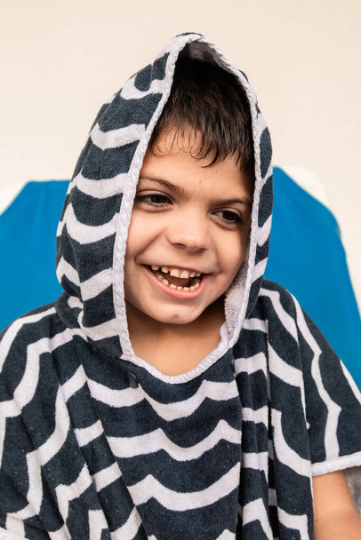 capelli castani ragazzo caucasico ispanico sorride felicemente con un accappatoio a righe blu e grigio e con cappuccio. foto ritratto - Foto, immagini