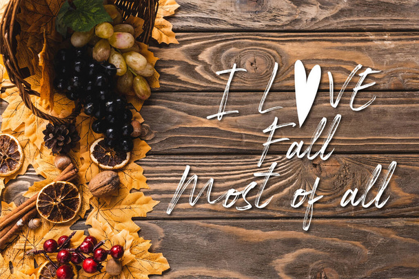 top view of autumnal harvest scattered from basket on foliage near i love fall most of all lettering on wooden background - Foto, Imagen