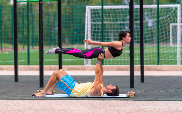 Young couple performing acrobatic yoga outdoors with the young man holding up and supporting the woman midair in horizontal position outdoors at a gym in a health and fitness or active lifestyle concept - Foto, Imagen