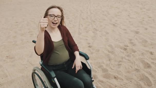 Młoda kobieta na wózku inwalidzkim na piaszczystym brzegu rzeki pokazuje kciuk w górę - Materiał filmowy, wideo