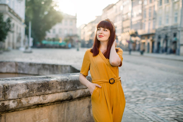 Portrait of beautiful smiling pretty woman in fashionable yellow clothes, posing to camera on a street of ancient European city. People, city portrait and lifestyle concept - 写真・画像