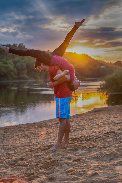 Young couple performing acrobatics yoga outdoors on a sandy beach at sunset or sunrise with reflections on the tranquil water in a health and fitness or active lifestyle concept - Фото, зображення