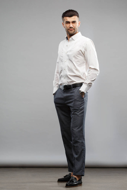 An elegant man in casual business clothes is standing tall with his hands in his pockets on a gray background. - Photo, Image