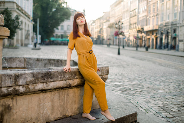 Horizontal shot of young beautiful Caucasian woman in fashionable yellow overalls, posing to camera with smile in urban environment, standing near ancient stone fountain. City woman portrait - Фото, зображення
