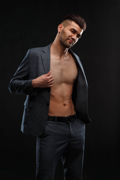 A young sexy man takes off his jacket worn over his naked body against a dark background. - Foto, Imagem