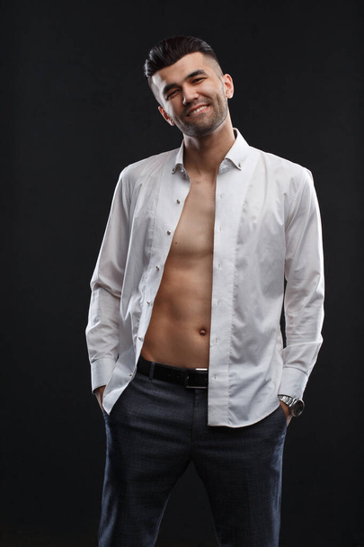 Charismatic attractive man in a unbuttoned white shirt hands in his trouser pocket isolated on a black background. - Foto, afbeelding