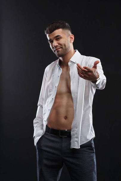 Charismatic attractive man in a unbuttoned white shirt stretched out his hand forward in a beckoning gesture isolated on a black background. - Photo, image