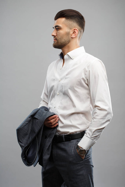 Young stylish man in a white shirt holds a suit jacket on his hand isolated on a gray background. - Foto, imagen