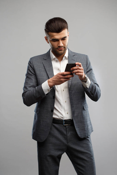 Stylish young man in an elegant suit holds a smartphone in his hands Isolated on a gray background. - Fotoğraf, Görsel
