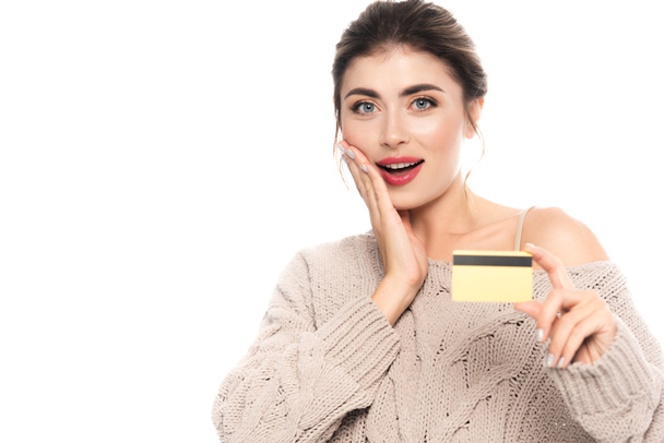 mujer emocionada en suéter de moda que muestra la tarjeta de crédito mientras mira la cámara aislada en blanco - Foto, Imagen