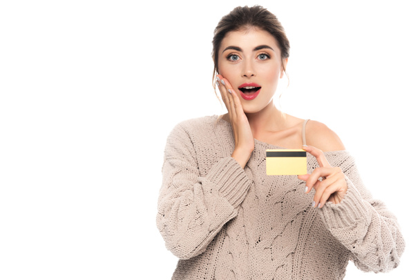 mujer conmocionada en suéter de punto tocando la cara mientras sostiene la tarjeta de crédito aislada en blanco - Foto, Imagen