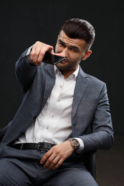 Young attractive man points forward with index finger holding a smartphone in this hand, isolated on a dark background. - Photo, Image