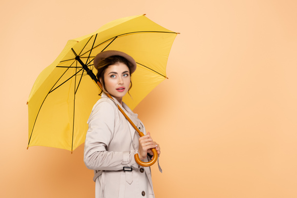 elegant woman in trench coat and beret looking away under yellow umbrella on peach - Фото, изображение