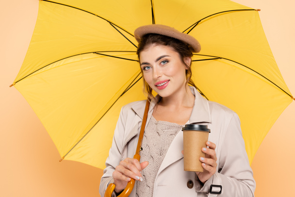 trendy woman in trench coat and beret holding coffee to go under yellow umbrella on peach - Photo, Image