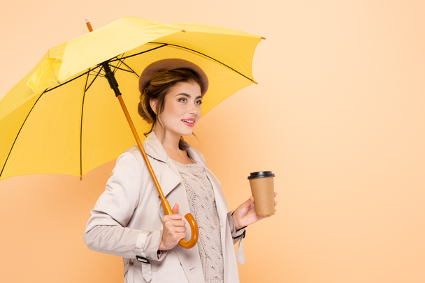 elegant woman in trench coat and beret holding paper cup under yellow umbrella on peach - Фото, изображение