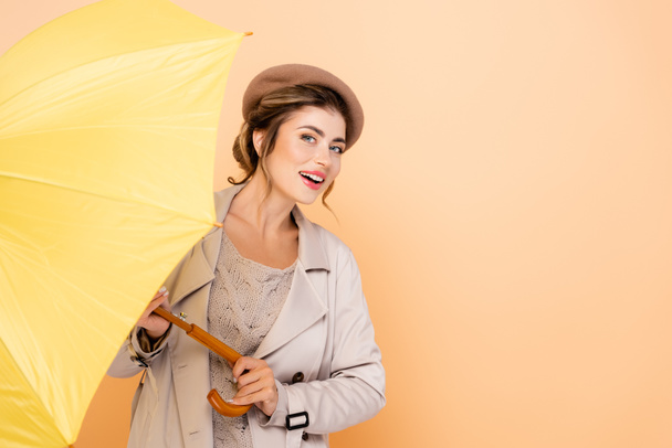 smyslná žena v módním plášti a baretu při pohledu do kamery, zatímco drží žlutý deštník na broskvi - Fotografie, Obrázek