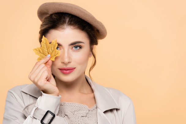 trendy woman in autumn outfit covering eye with yellow leaf while looking at camera isolated on peach - Photo, Image