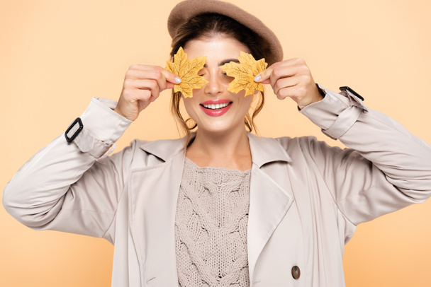mulher alegre em casaco de trincheira elegante e boina cobrindo olhos com folhas amarelas isoladas em pêssego - Foto, Imagem