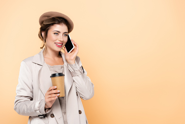 jovem mulher em roupa de outono elegante segurando café para ir enquanto conversa no smartphone isolado em pêssego - Foto, Imagem