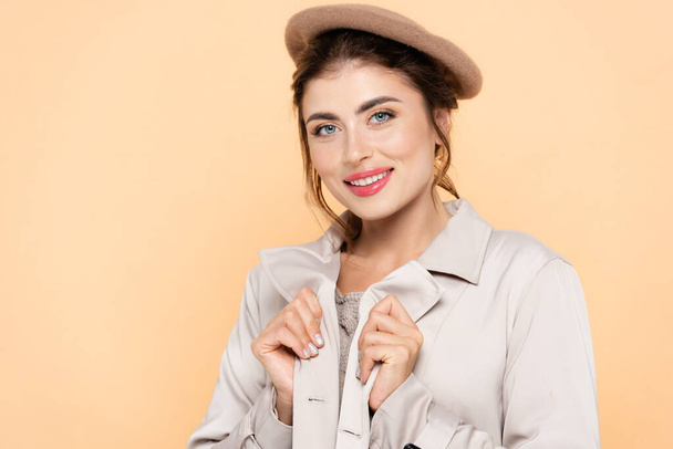 young woman in beret touching trench coat while looking at camera isolated on peach - Foto, afbeelding