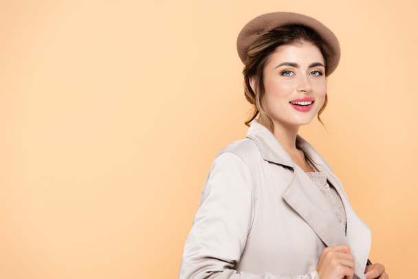 sensual woman in stylish beret and trench coat looking at camera while posing isolated on peach - 写真・画像