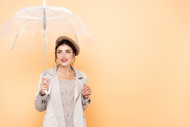 Trençkotlu ve bereli neşeli kadın şeftali rengi şemsiyenin altında başka tarafa bakıyor. - Fotoğraf, Görsel