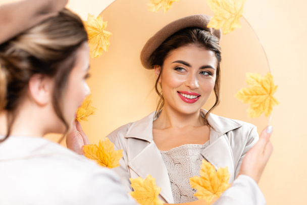 selective focus of young woman in trench coat and beret looking in mirror decorated with autumn leaves on peach - Foto, immagini