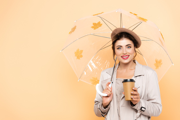 joyful woman in stylish autumn outfit holding coffee to go under umbrella with foliage on peach - Photo, Image