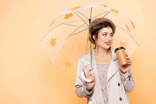 mujer de moda en boina y gabardina sosteniendo café para ir bajo paraguas con hojas de otoño en melocotón - Foto, Imagen