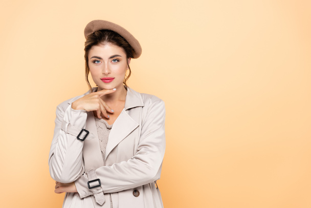 fashionable woman in trench coat and beret touching chin while looking at camera on peach - Photo, Image