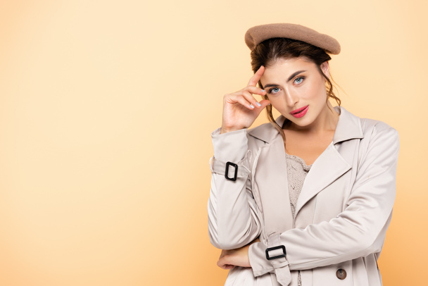 fashionable woman in trench coat and beret touching forehead while looking at camera on peach - Fotoğraf, Görsel
