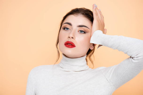 stylish woman in turtleneck looking away while touching face isolated on peach  - Foto, Bild