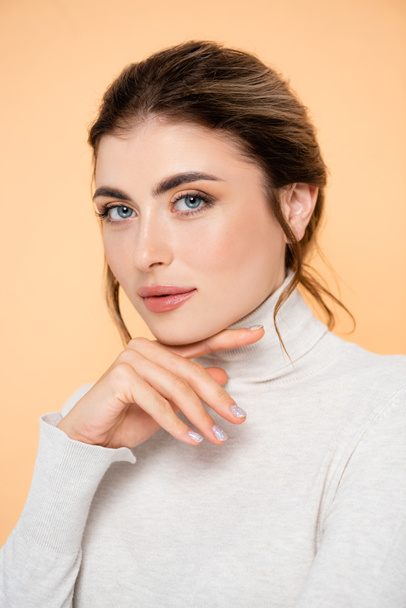 stylish woman in turtleneck touching chin and looking at camera isolated on peach  - Фото, изображение