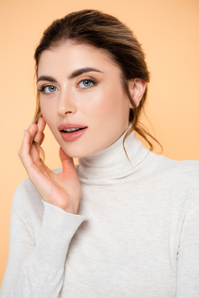 young woman in turtleneck looking at camera while touching face isolated on peach  - Foto, afbeelding