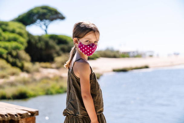 retrato de uma menina loira com olhos azuis vestindo uma máscara facial em férias em uma praia com pinheiros com um rosto sério em um vestido verde no meio de uma pandemia de coronavírus covid19 - Foto, Imagem