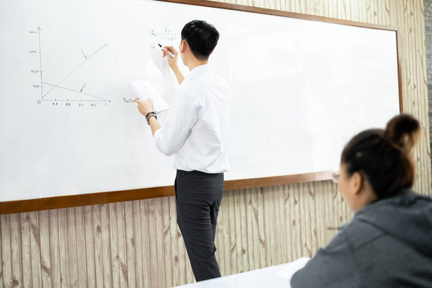 An Asian male teacher in a classroom near the whiteboard is writing and teaching a lesson with a special lecture in a class column. Math teacher explains arithmetic sums to university students. - Zdjęcie, obraz