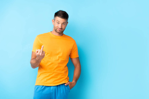 άνδρας σε t-shirt στέκεται με το χέρι στην τσέπη και δείχνει το μεσαίο δάχτυλο στο μπλε - Φωτογραφία, εικόνα