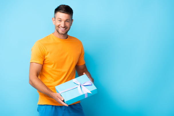 happy man in t-shirt holding wrapped gift box on blue - Photo, Image
