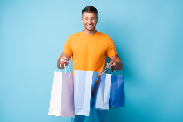 cheerful man in t-shirt holding shopping bags on blue - Foto, imagen