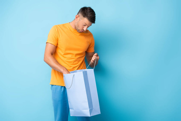 surprised man in t-shirt looking at shopping bag on blue - Foto, Imagem
