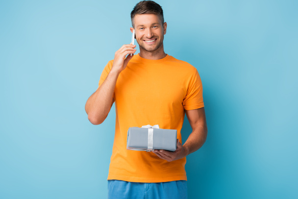 Χαρούμενος άνδρας με t-shirt κρατώντας κουτί δώρου και μιλώντας στο smartphone σε μπλε - Φωτογραφία, εικόνα