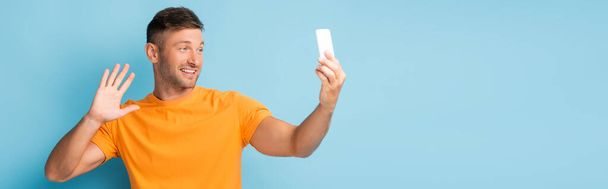 άνδρας σε t-shirt κρατώντας smartphone και τη λήψη selfie, ενώ κυματίζει το χέρι σε μπλε, banner - Φωτογραφία, εικόνα
