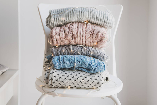 Pile of warm woolen sweaters wrapped in decorative Christmas lights on white chair. Handmade knitwear. Warm clothing - Photo, Image