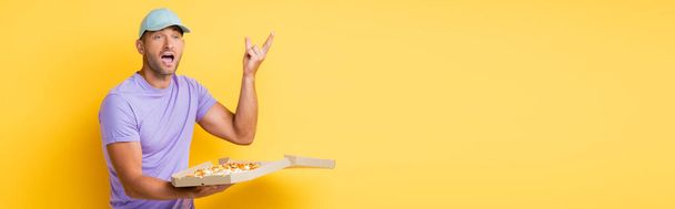 homem de chapéu azul gesticulando enquanto segurando caixa com pizza saborosa no amarelo, banner - Foto, Imagem