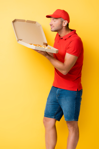 zadowolony dostawca w czerwonym wieczku pachnący smaczną pizzą w pudełku kartonowym na żółto - Zdjęcie, obraz
